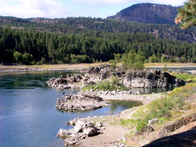 [A rocky shorelines sticks out into the river in an irregular fashion. Plenty of evergreens on the far shore of the river with a mountain in the far distance on the right.]